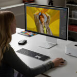 Person sits at a white desk editing a photo of a person on a large monitor, with a color swatch nearby in a bright studio setup.