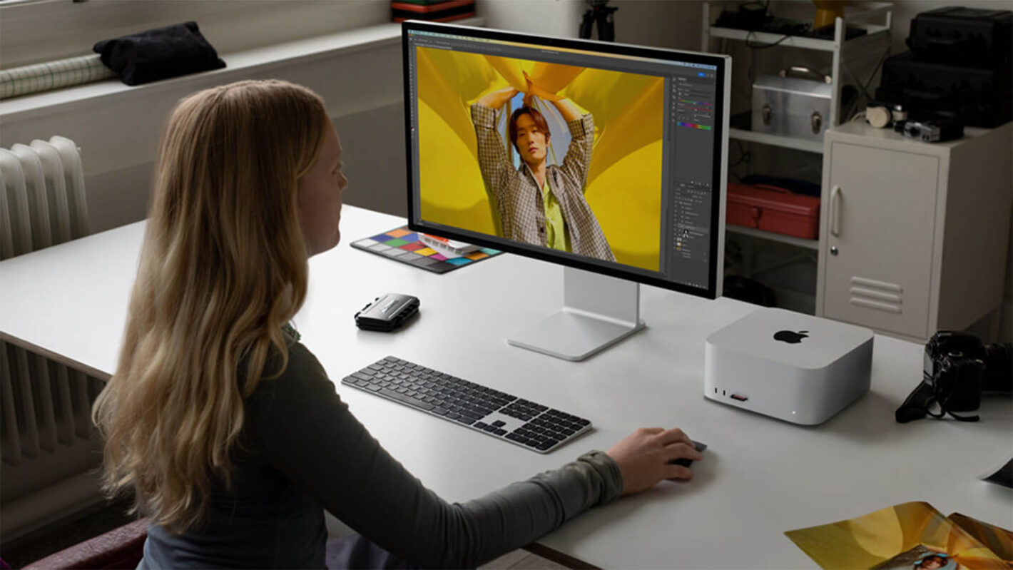Person sits at a white desk editing a photo of a person on a large monitor, with a color swatch nearby in a bright studio setup.