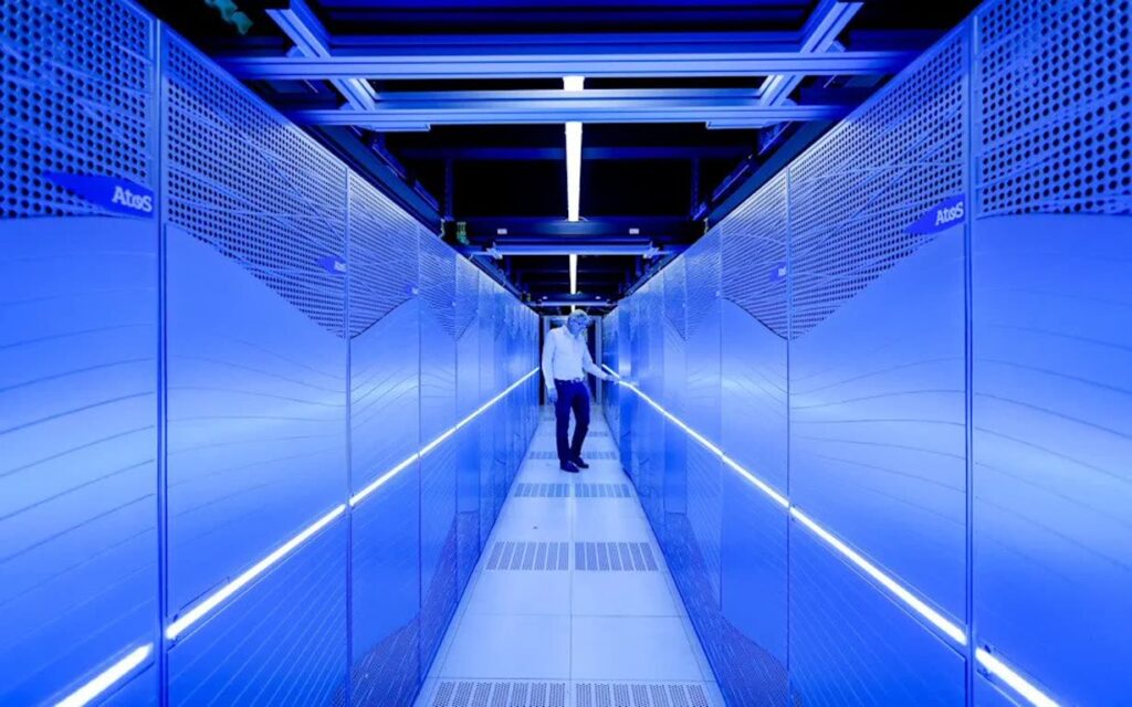 Man in a white shirt walks down a narrow blue-lit data center aisle between server racks.