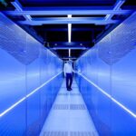 Man in a white shirt walks down a narrow blue-lit data center aisle between server racks.