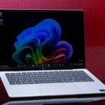 Open silver laptop displaying a vibrant abstract blue-to-green wallpaper on a glossy black surface against a red and purple checkered background.