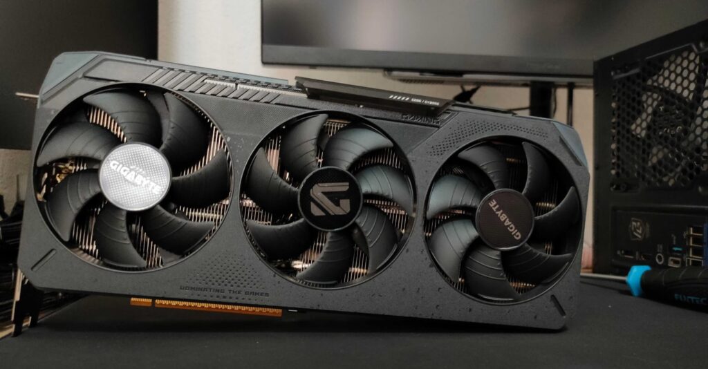 Triple‑fan graphics card from Gigabyte installed in a PC, visible heatsink and case side panel in the background, slogan 'Dominating the Games' on the card.